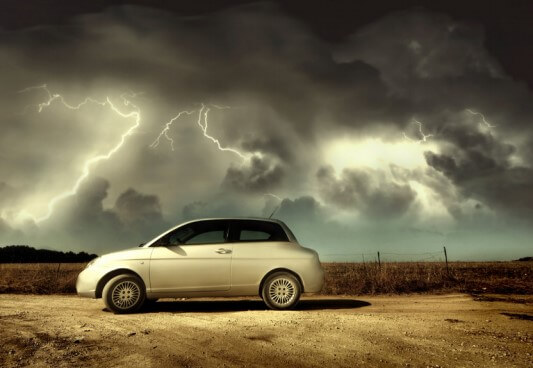 Na imagem, há um carro estacionado e uma tempestade no céu: muitas nuvens escuras e relâmpagos.