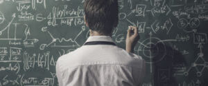 Professor olhando para lousa verde com fórmulas matemáticas escritas em giz branco