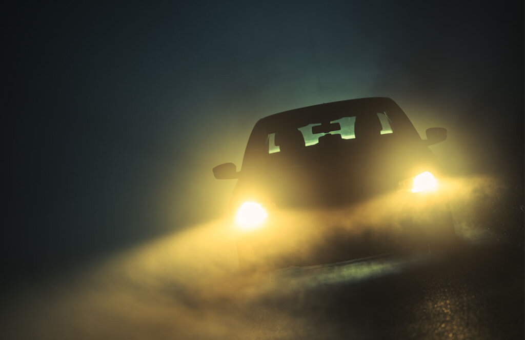 A imagem mostra um carro no escuro, com faróis acesos e a reflexão da luz da neblina, que é um coloide do tipo aerossol