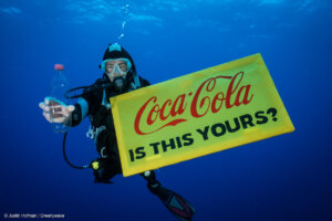 Um mergulhador do Greenpeace segura uma faixa com os dizeres "Coca-Cola é sua?" e uma garrafa de Coca-Cola encontrada à deriva na lixeira. The Great Pacific Garbage Patch (ou "Grande Porção de Lixo do Pacífico" traduzido para o português) é uma grande mistura de plásticos e microplásticos, agora com o dobro do tamanho do Texas, no meio do Oceano Pacífico Norte.