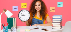 Menina estudando em uma mesa, com objetos como livros, caderno e lápis. Ela segura um relógio e há post its com a palavra vestibular no fundo.