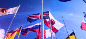 Bandeiras de diversos países hasteadas, com céu azul ao fundo.