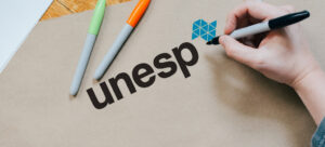 Imagem mostra a palavra "Unesp" sendo escrita e o logo a universidade em uma folha de papel, ao lado de canetinhas coloridas