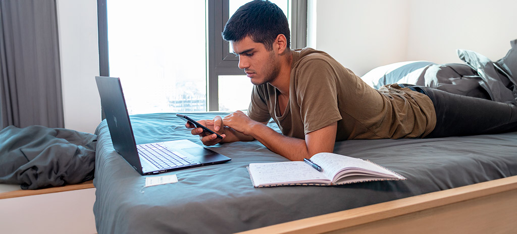 Garoto deitado de bruços na cama mexendo no celular com livro e notebook abertos como sinônimo de procrastinação