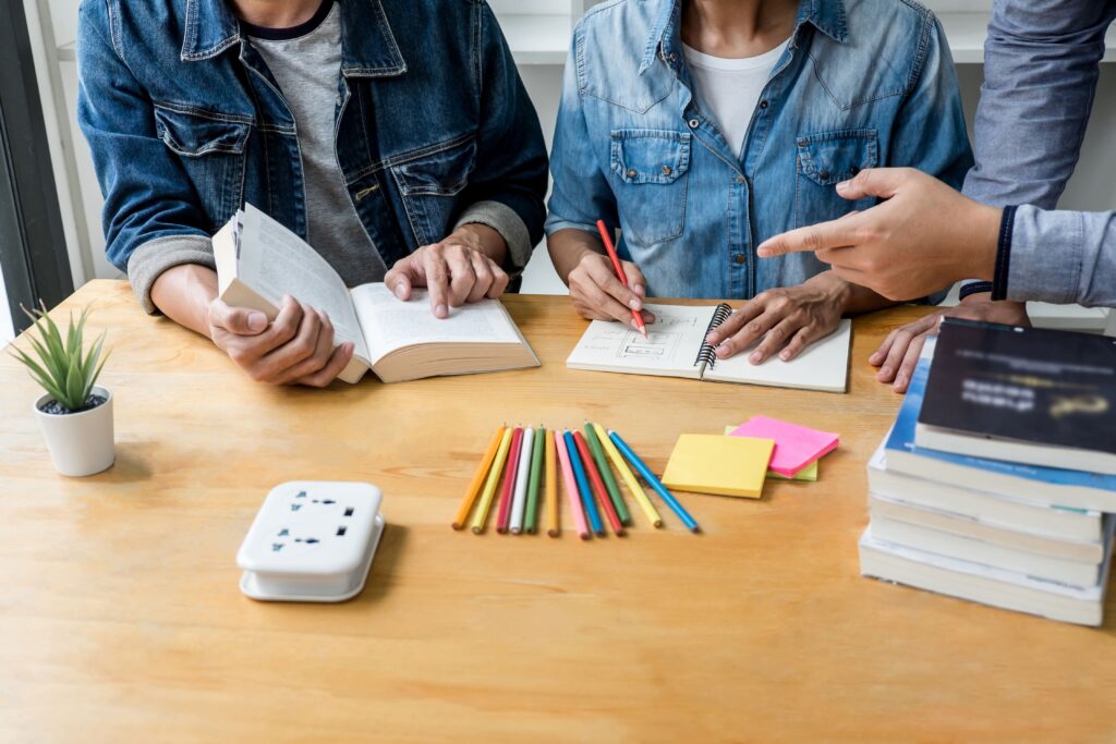 Dois estudantes, um lendo um livro e outro escrevendo em um caderno, estão estudando juntos em uma mesa com lápis coloridos e pilhas de livros