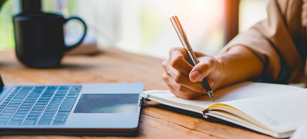 Uma pessoa está escrevendo em um caderno ao lado de um laptop e uma caneca em uma mesa de madeira
