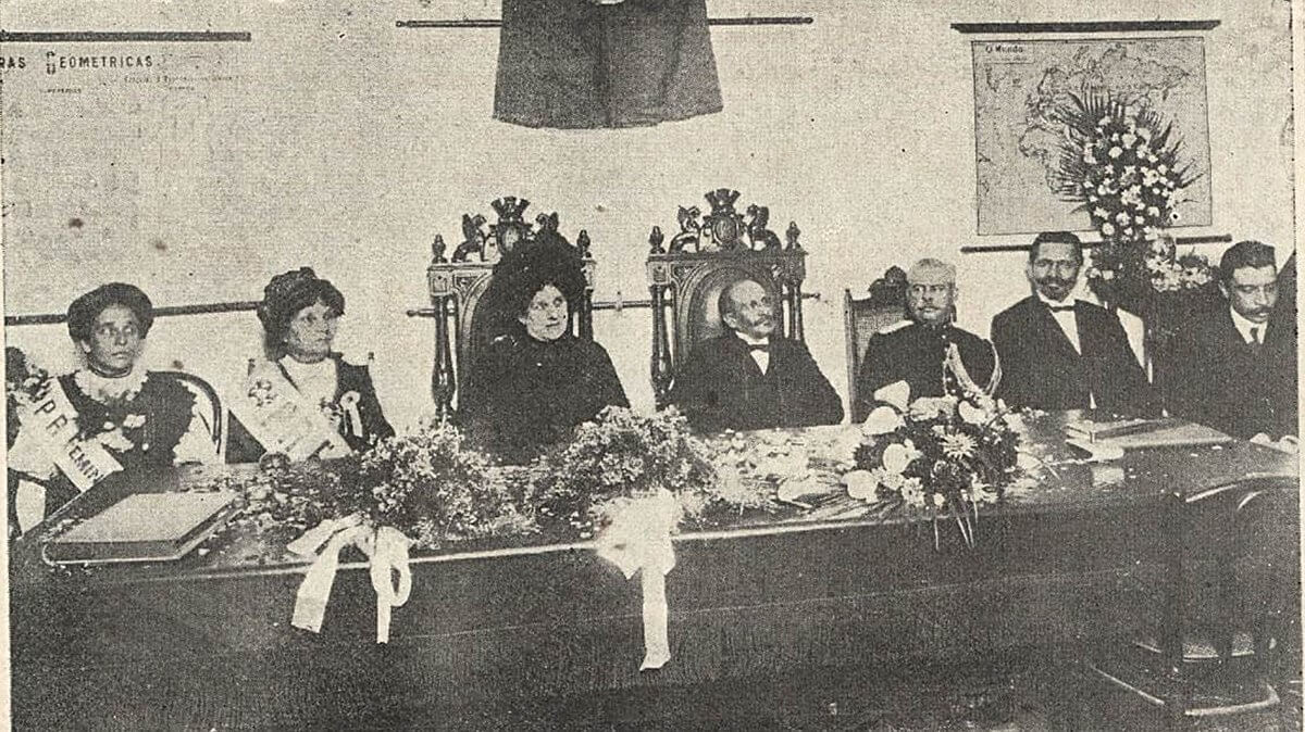Foto em preto e branco do momento de inauguração da Escola de Ciências e Artes Orsina Fonseca. Na mesa, estão sentados o presidente Hermes da Fonseca, sua esposa e Leolinda Daltro.