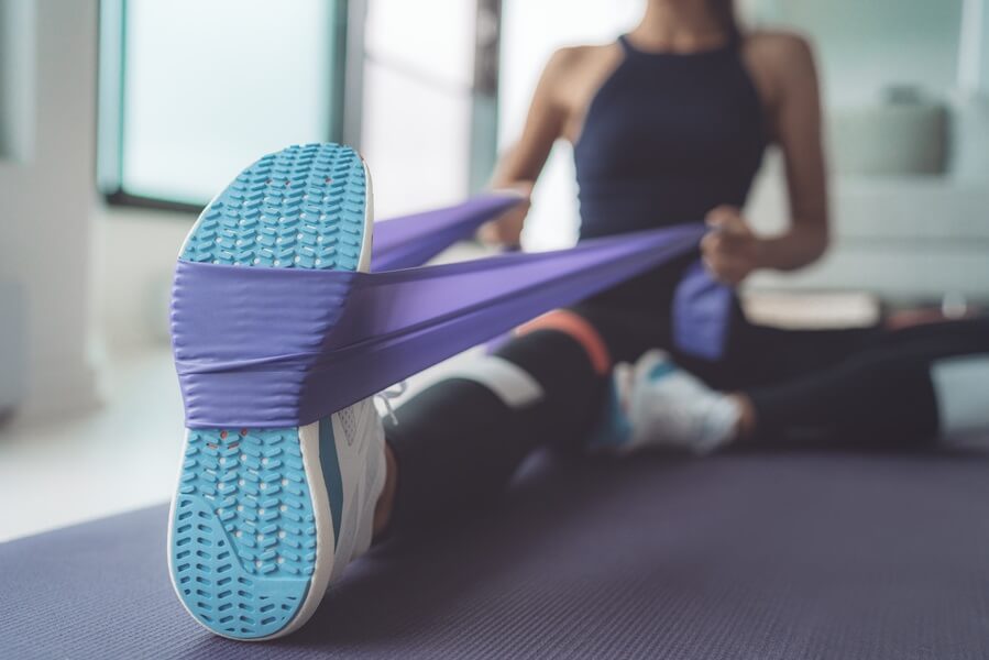 Mulher em academia com roupa de ginástica sentada no chão e com elástico embaixo do pé puxando as duas pontas com as mãos