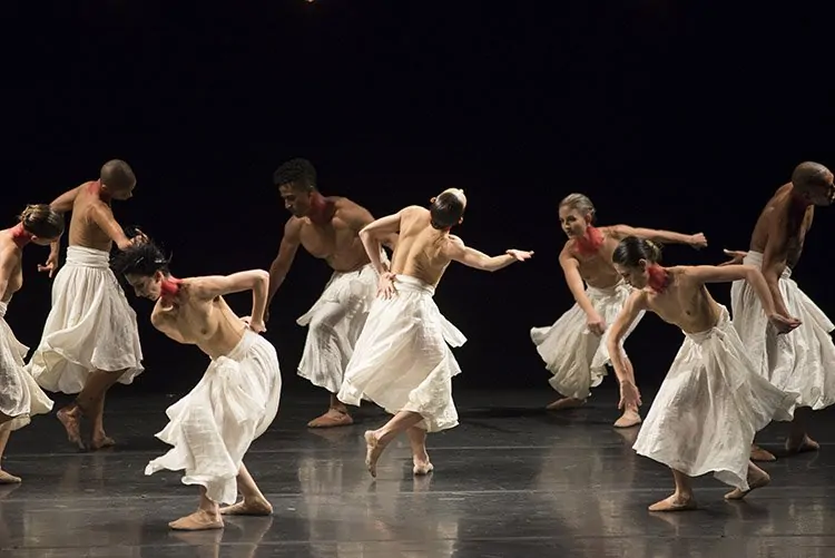 Imagem referenciada em questão de Literatura no Enem. A foto mostra o espetáculo de Bailarinos em trajes brancos fluídos executam uma dança contemporânea num palco escuro.