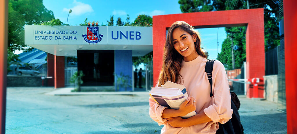 Mulher jovem com mochila segurando livros na frente da portaria da Uneb após ser aprovada no vestibular da Uneb