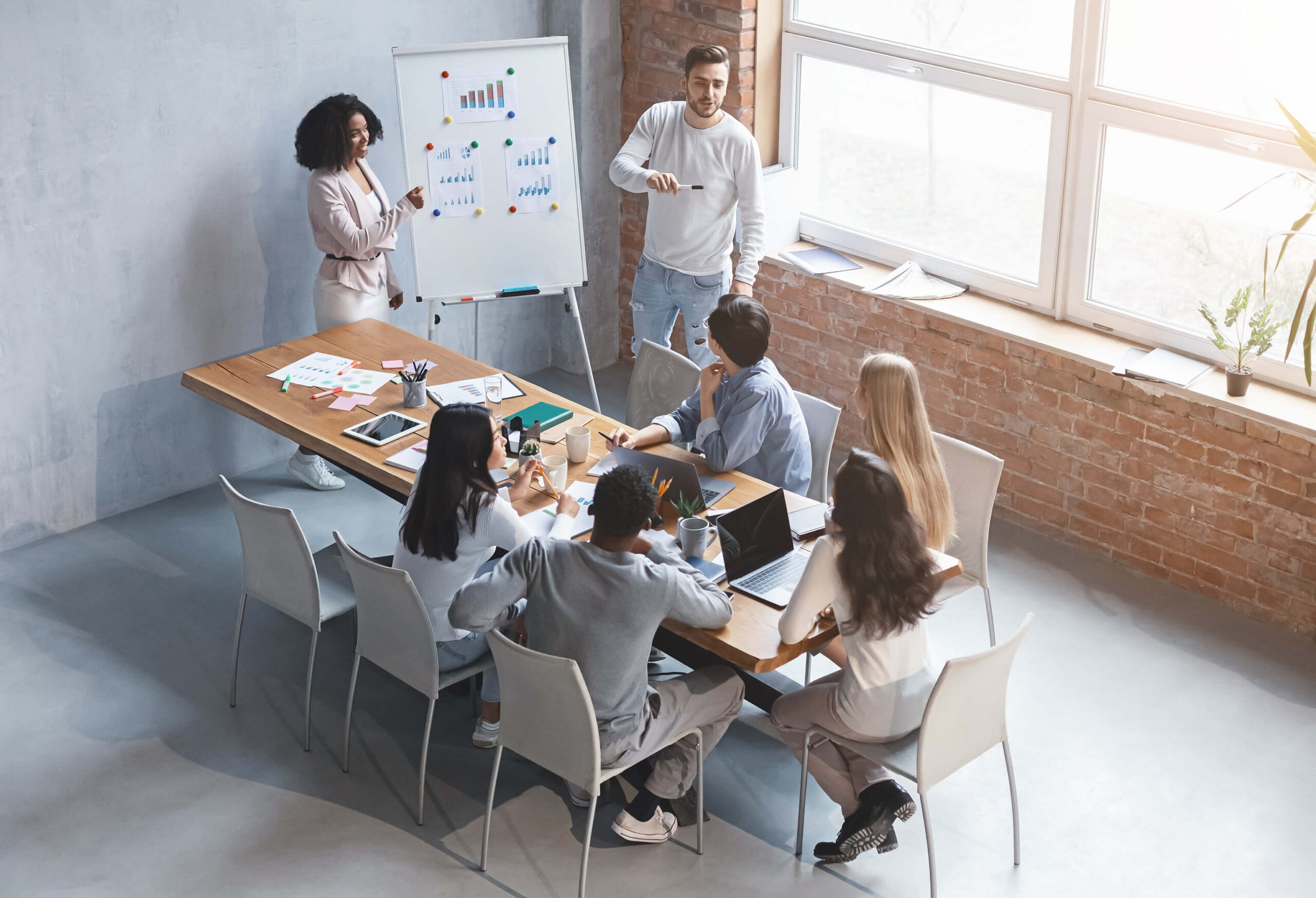 Equipe de marketing discute estratégias em uma reunião, com dois apresentadores explicando gráficos em um quadro branco