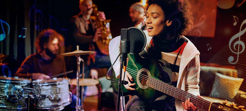 Aluna do curso de Música em estúdio de gravação tocando violão e cantando em microfone. Ao fundo, baterista e saxofonista tocando instrumentos