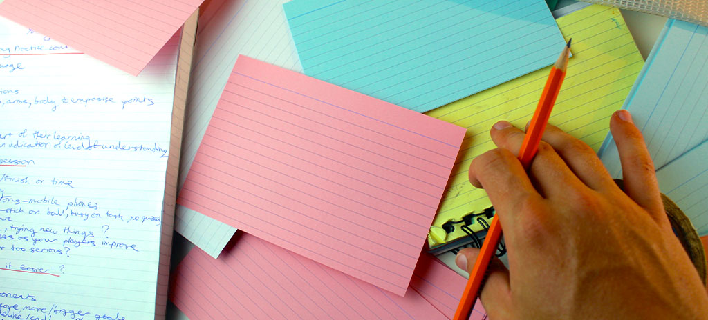 Conjunto de flashcards coloridos espalhados sobre uma mesa, acompanhados de uma folha de caderno com anotações manuscritas e uma mão segurando um lápis laranja