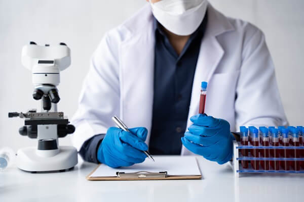 Biomédico de jaleco branco, máscara e luvas azuis analisando amostras de sangue em tubos com um microscópio ao lado