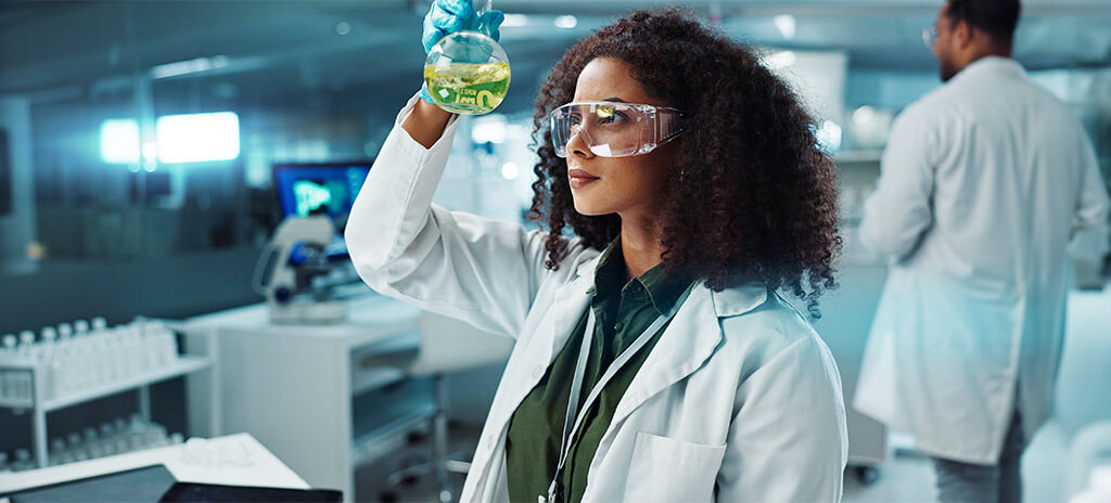 Aluna da faculdade de engenharia química em laboratório com jaleco, luvas e óculos de proteção segurando vidro com substância líquida amarela