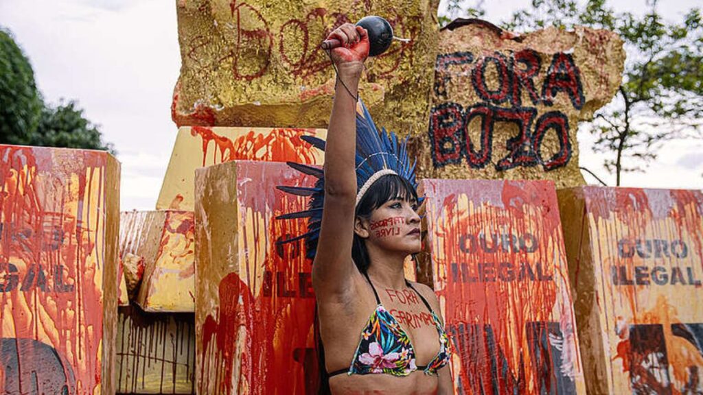 Mulher indígena protestando contra o garimpo ilegal durante o Dia Internacional dos Povos Indígenas, com cartazes pintados de vermelho ao fundo