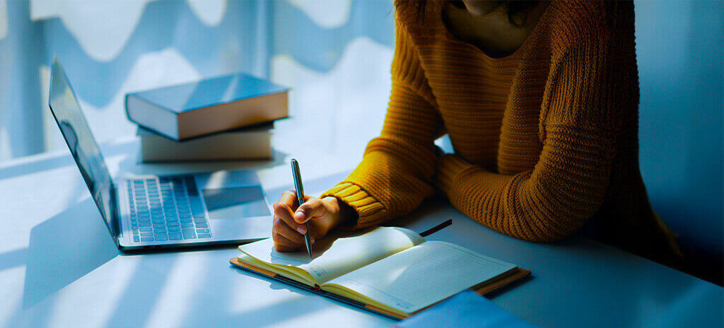 Mulher de suéter amarelo escrevendo metas de estudo com caneta em um agenda. Ela está apoiada em uma escrivaninha, com um notebook e livros ao lado.