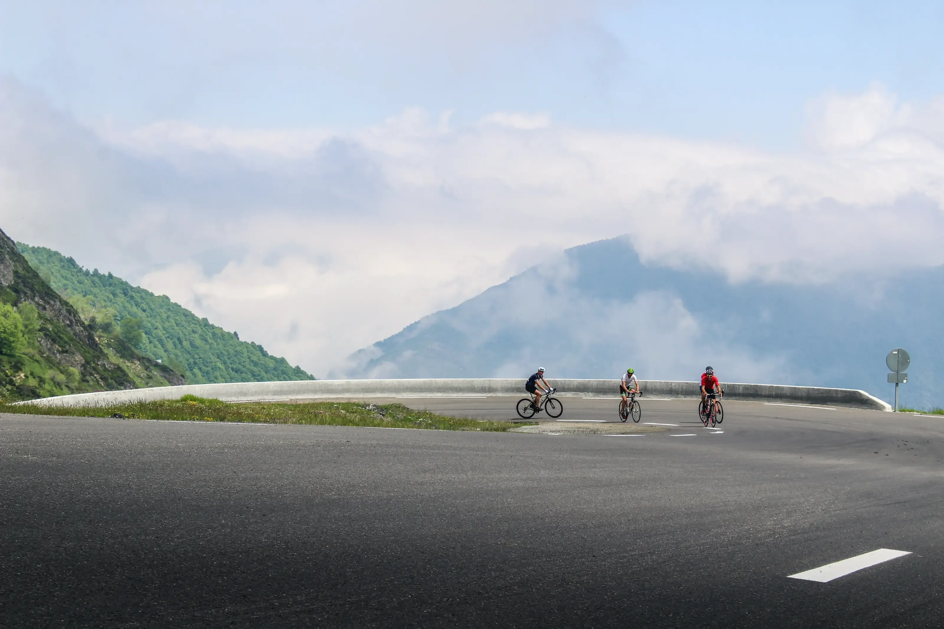 Quatre cyclistes négocient un grand virage en lacet sur une route de montagne, face à des sommets verdoyants et brumeux.