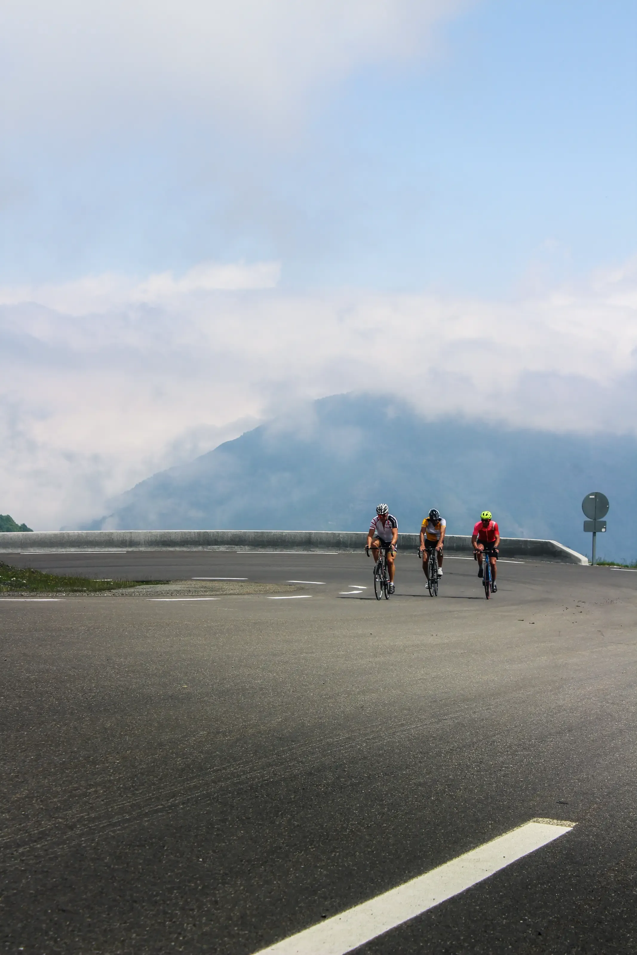 Trois cyclistes négocient un grand virage en lacet sur une route de montagne, face à un sommet brumeux sous un ciel nuageux.