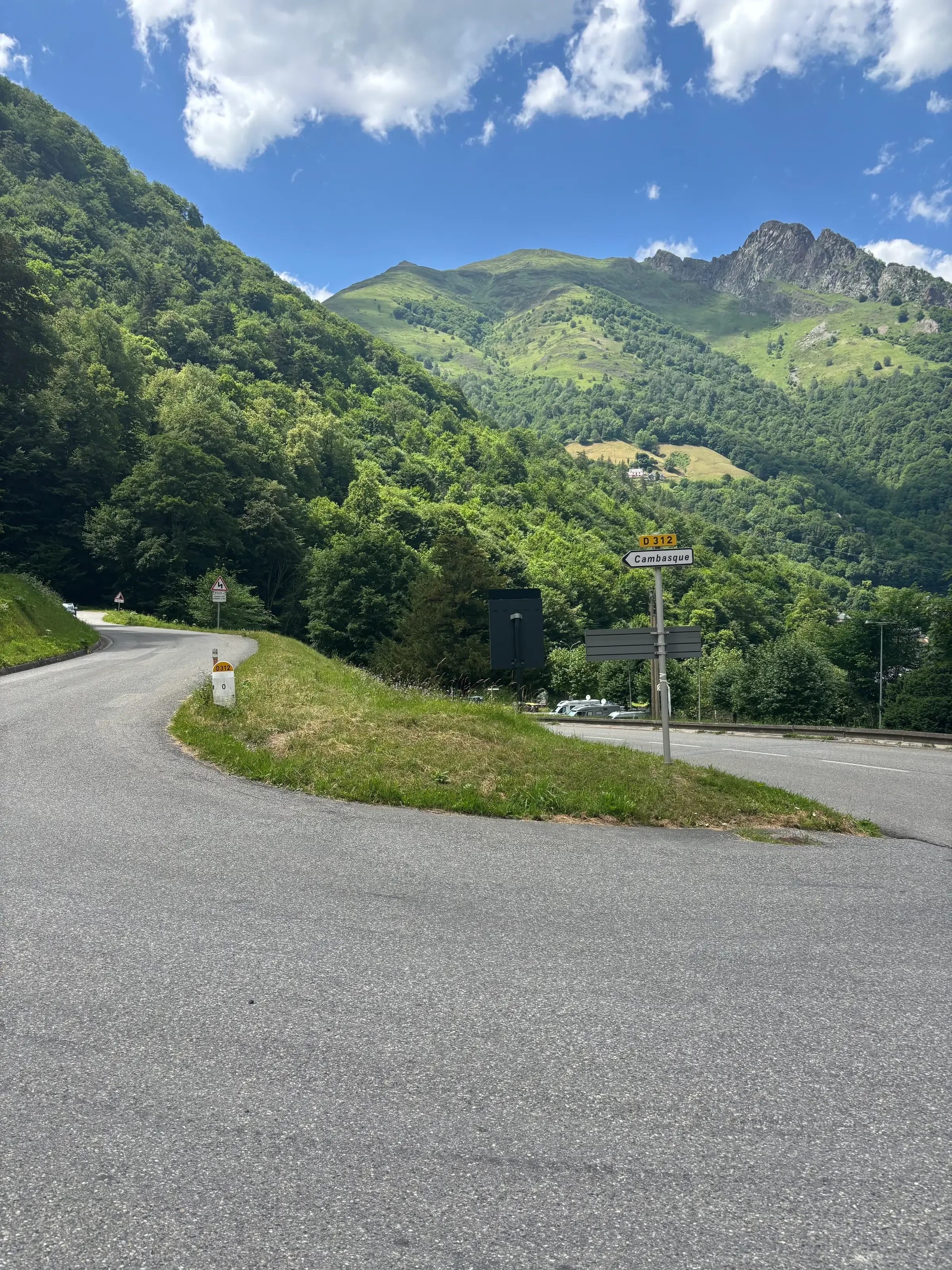 Une route de montagne goudronnée forme un virage près de panneaux de signalisation, face à de grands sommets verdoyants sous un ciel bleu parsemé de nuages blancs.