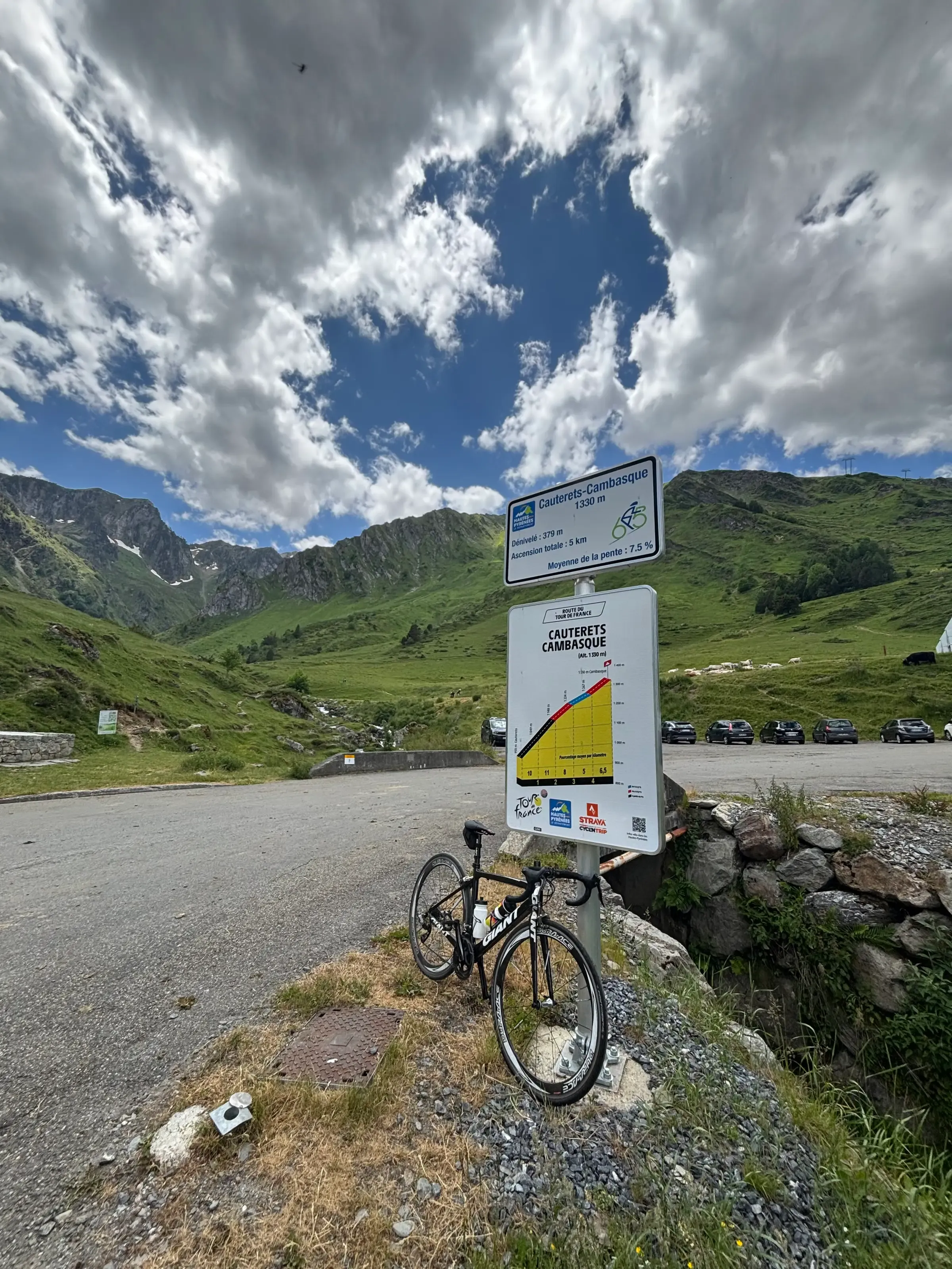 Un vélo de route est appuyé contre des panneaux d'information de Cauterets-Cambasque, face à de grands sommets verdoyants sous un ciel nuageux.