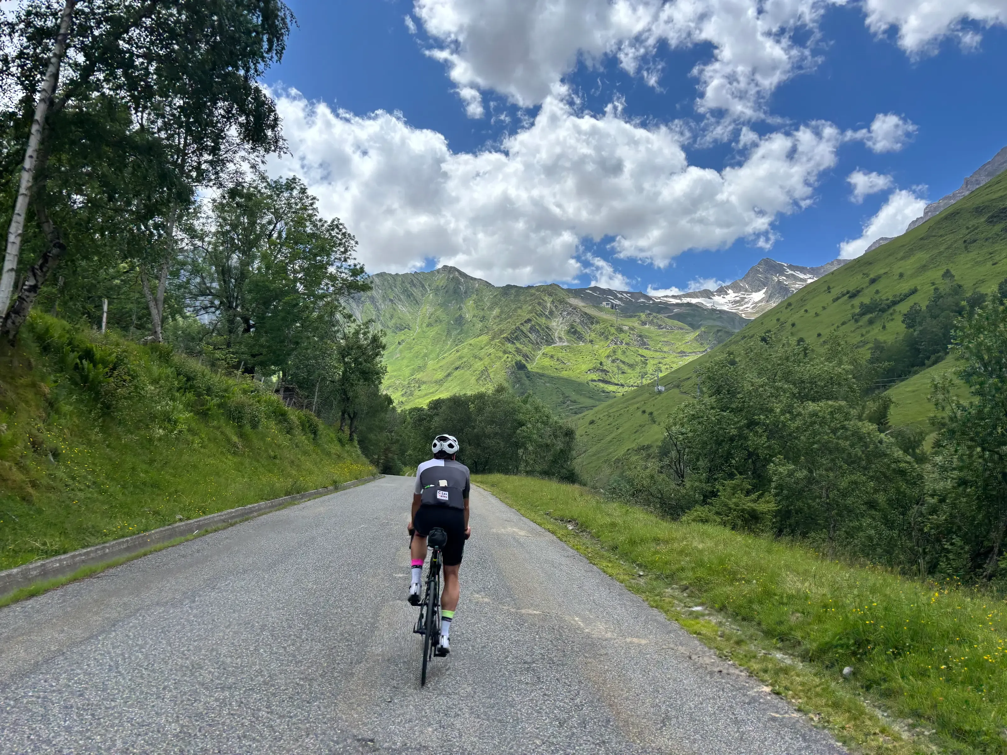 Un cycliste vu de dos gravit une route de montagne ensoleillée, face à de grands sommets verdoyants sous un ciel bleu parsemé de nuages blancs.