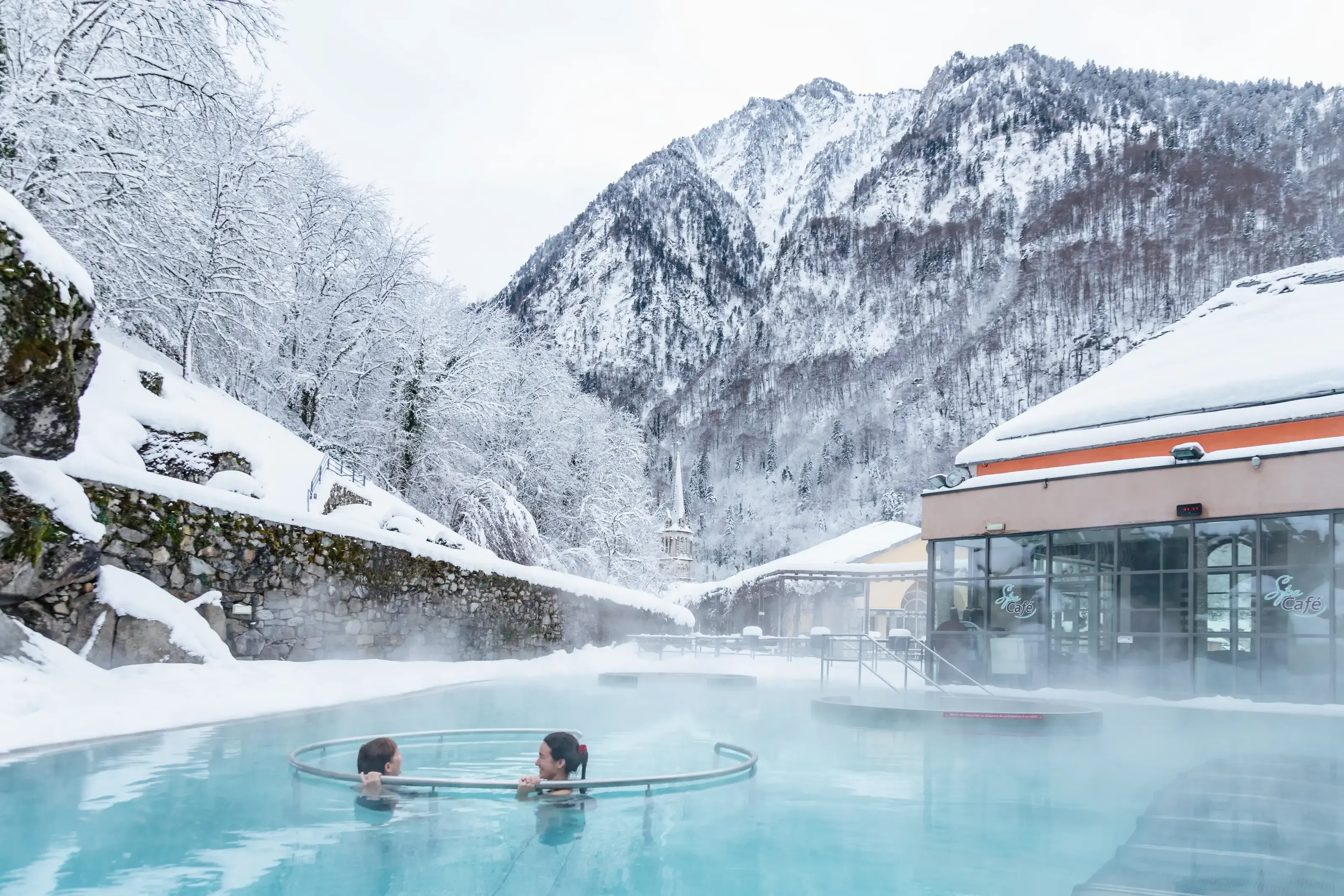Deux femmes profitants des eaux chaudes des Bains du Rocher alors que la neige entoure les montagnes.