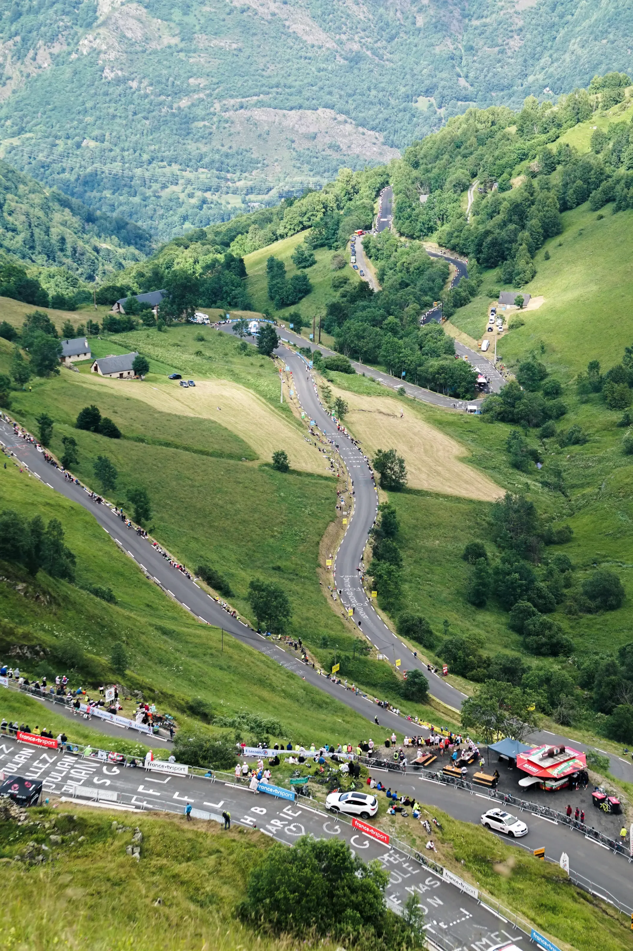 Une vue plongeante à angle vertical d'une route de montagne aux nombreux virages en lacets, bondée de spectateurs en bas et serpentant à travers des pentes verdoyantes.