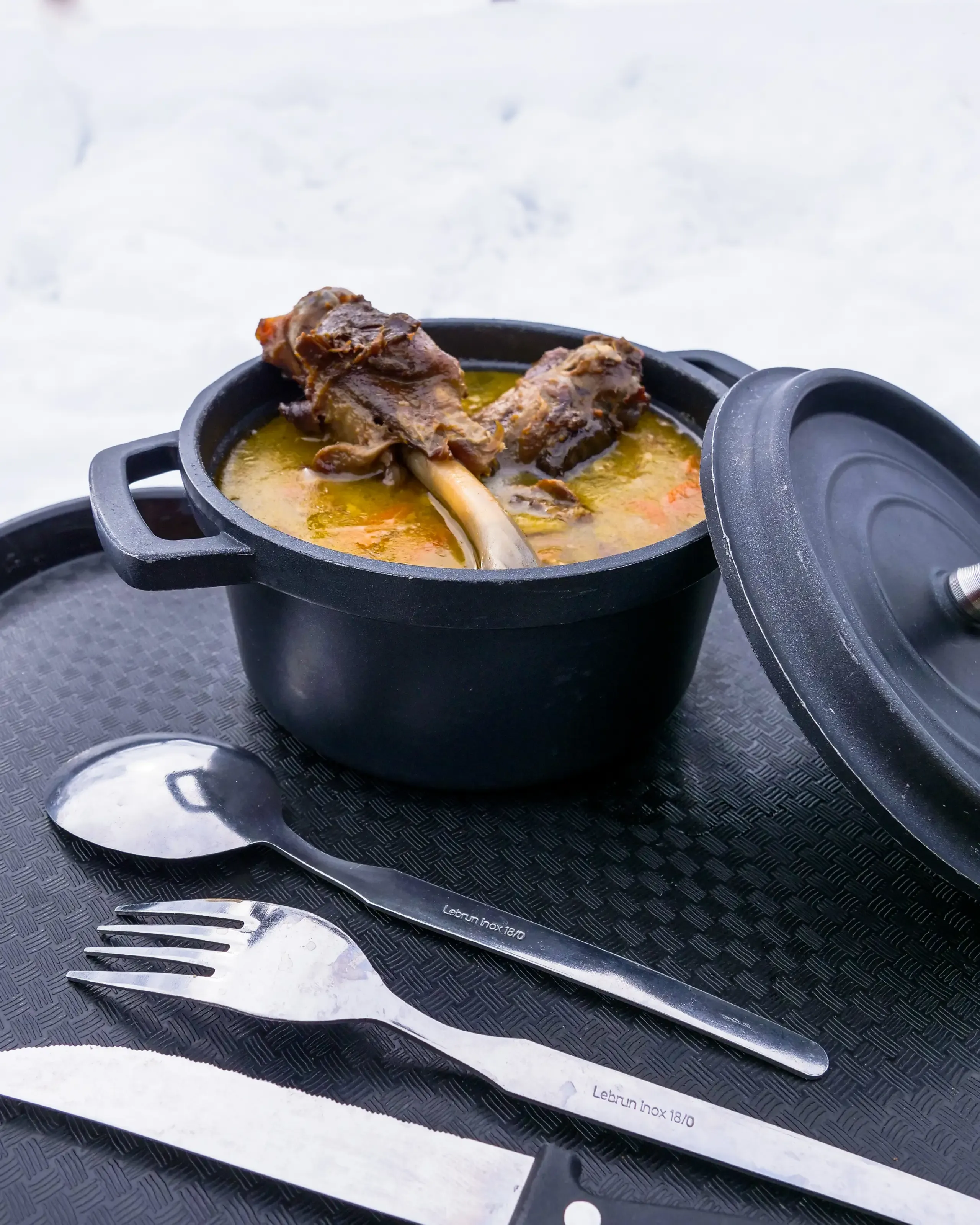 Garbure au confit de canard servie dans une cocotte en fonte noire sur un plateau, avec couverts, devant un fond de neige