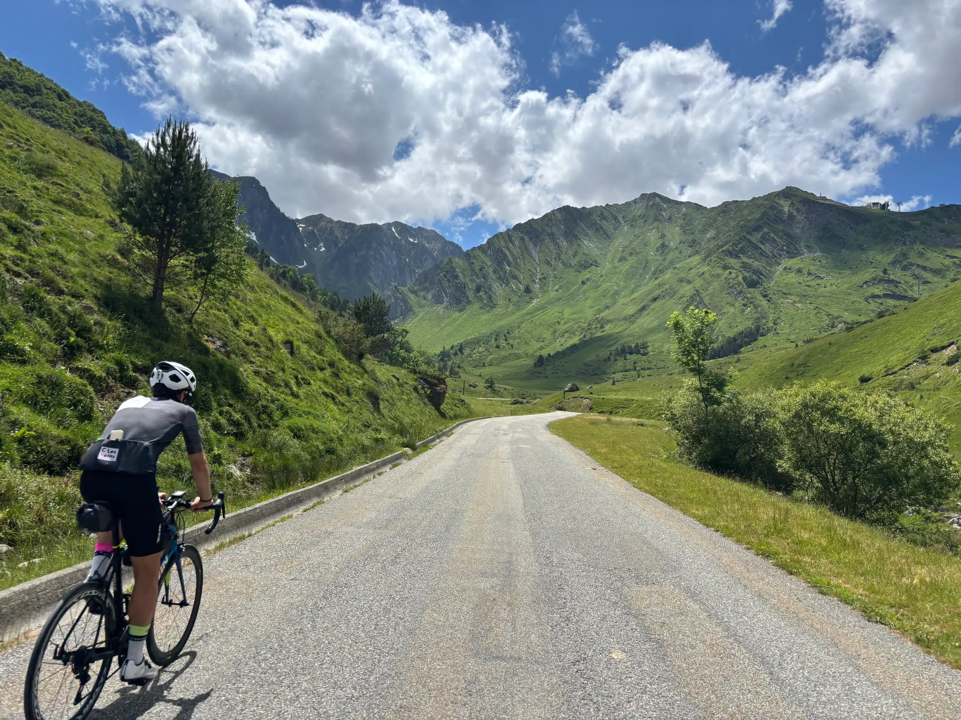 Un cycliste vu de dos roule sur une route de montagne ensoleillée, face à de grands sommets verdoyants sous un ciel bleu parsemé de nuages blancs.