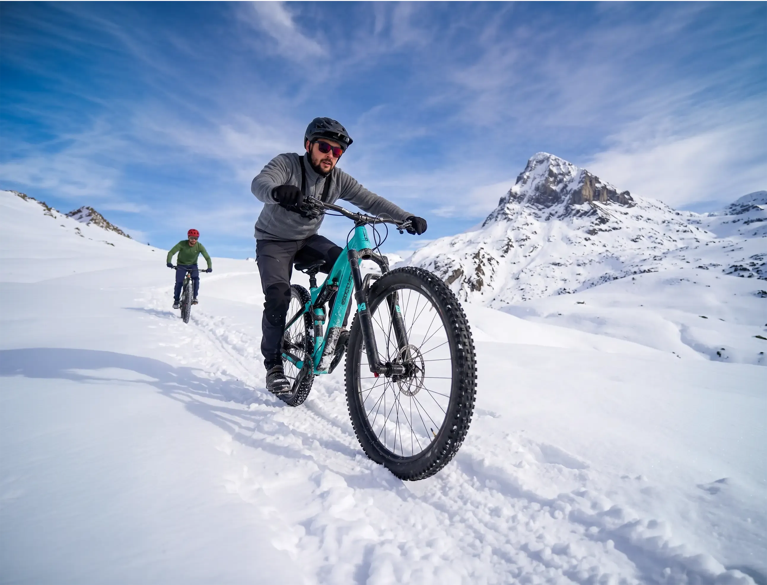 Deux vététistes descendant un sentier enneigé à VTT, face à un sommet rocheux sous un ciel bleu