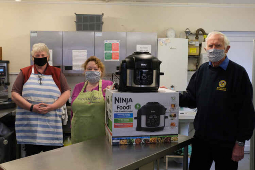 Karen Taylor, founder of the Nourish organisation in Axminster, and her assistant Mary Darlow, receives a pressure cooker from Rotarian Peter Creek at the Axminster Guildhall kitchen where the meals are provided