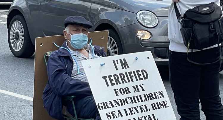 Pat sits on a chair in the middle of the road with two signs reading "I'm terrified for my grandchildren, sea level rise, coastal flooding, desertification, water shortage, wildfires, hurricanes" / Photo credit: @TeddingtonMill1