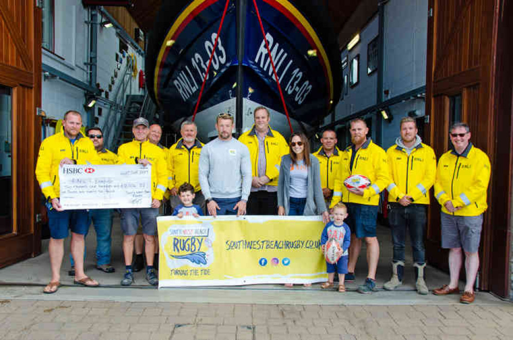 Holding Cheque, L – R, Roy Stott, Exmouth RNLI Deputy Coxswain; Steve Hockings-Thompson, Exmouth RNLI Coxswain.  Holding banner in front of Exmouth RNLI Crew Members, Matt and Sarah Jess and their two boys Ted and Alfie. Image : John Thorogood / Exmouth