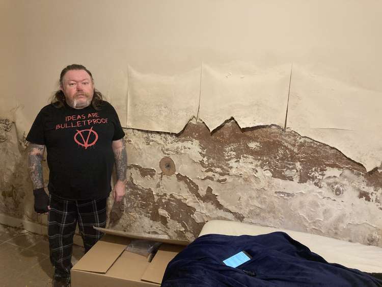 Michael Brady standing next to his bedroom wall where wallpaper has rotted and peeled off as a result of damp and mould caused by a leak. (Credit: Lisa Haseldine)