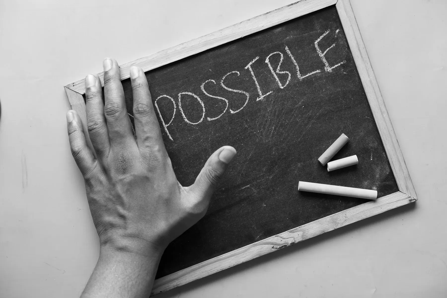 A hand resting on a small framed chalkboard with the word POSSIBLE written on it, two sticks of chalk lying beside it.