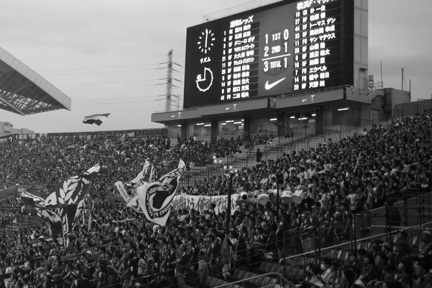 A stadium scoreboard showing both line-ups and a 3-1 score above a packed stand of supporters waving flags, in black and white.