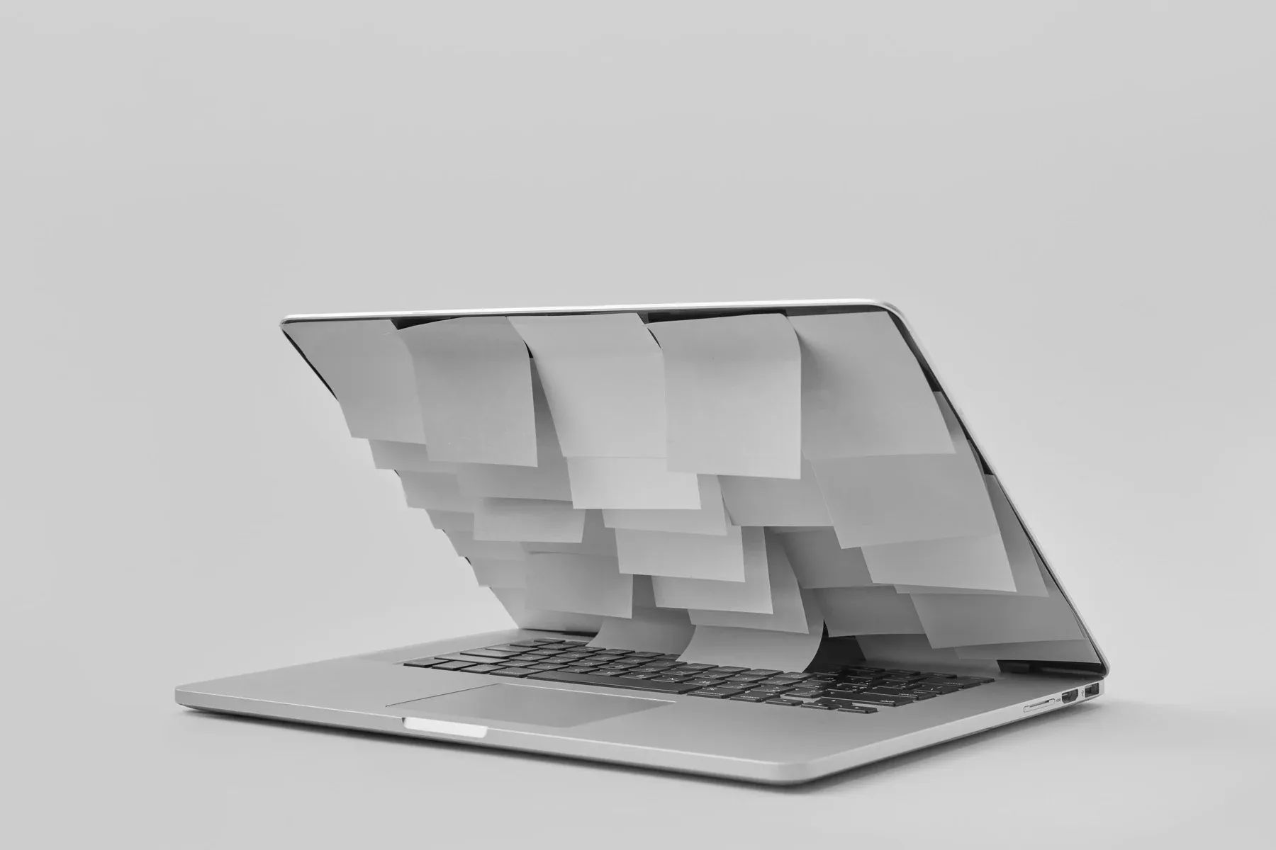An open laptop with sheets of blank paper hanging in overlapping rows from the top of its screen, on a plain grey background, in black and white.