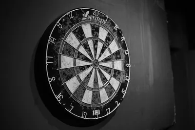 A well-used dartboard mounted on a dark wall, its numbered ring and scoring beds lit from one side.
