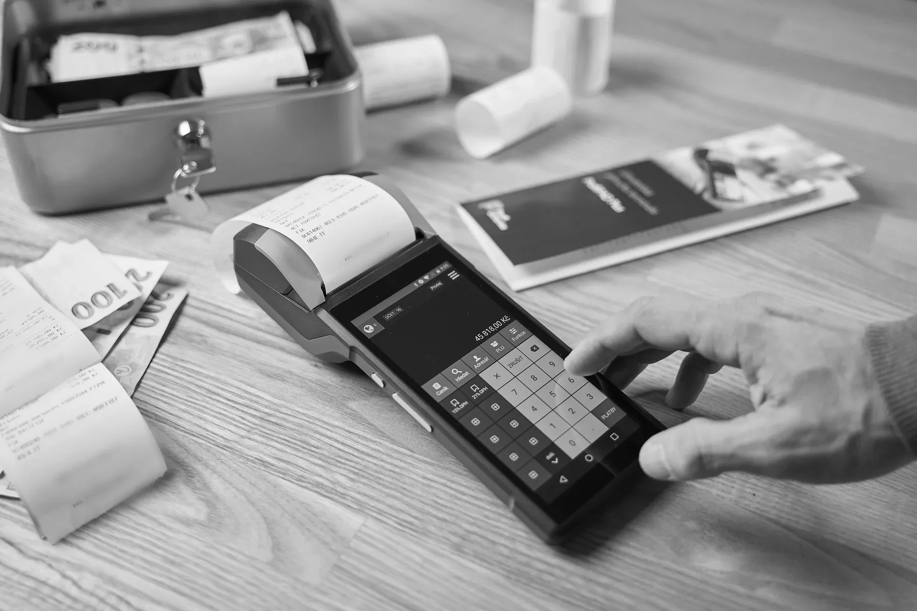 A hand entering an amount on a card payment terminal, a printed receipt curling from its printer, with a cash tin, banknotes and used receipts on the counter around it.