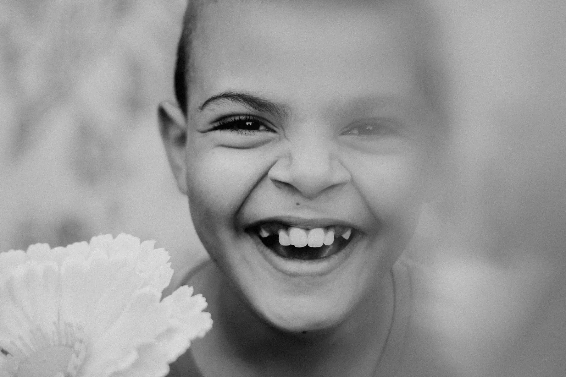 A young child laughing, photographed close up in black and white, with a pale flower held up beside their face.