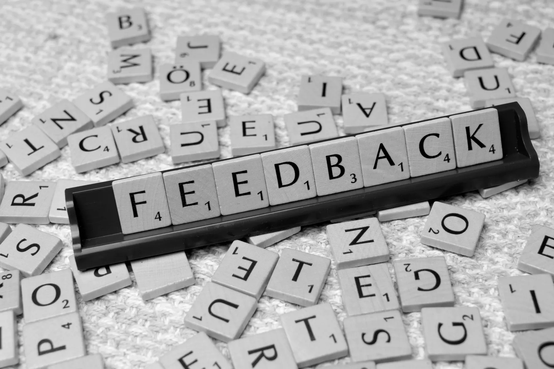 Scrabble-style letter tiles spelling the word FEEDBACK on a tile rack, with loose letter tiles scattered face up around it.
