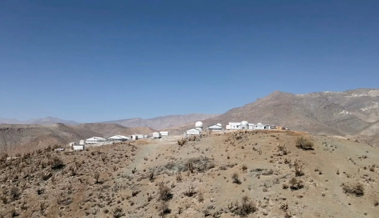 The observatory's white buildings and domes along a dry ridge, mountains behind, under a clear sky