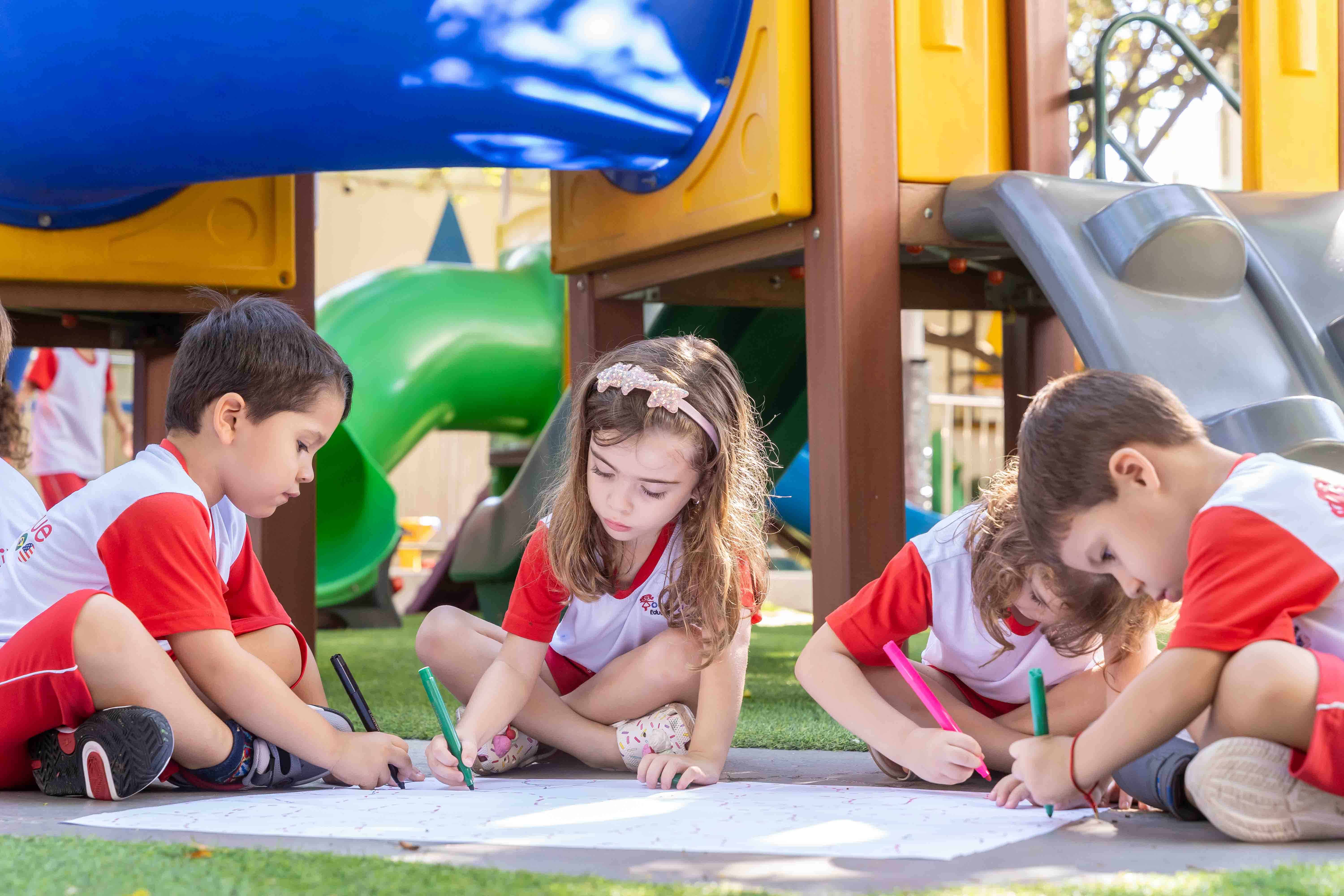 Três crianças desenhando com giz de cera sentadas na grama do playground