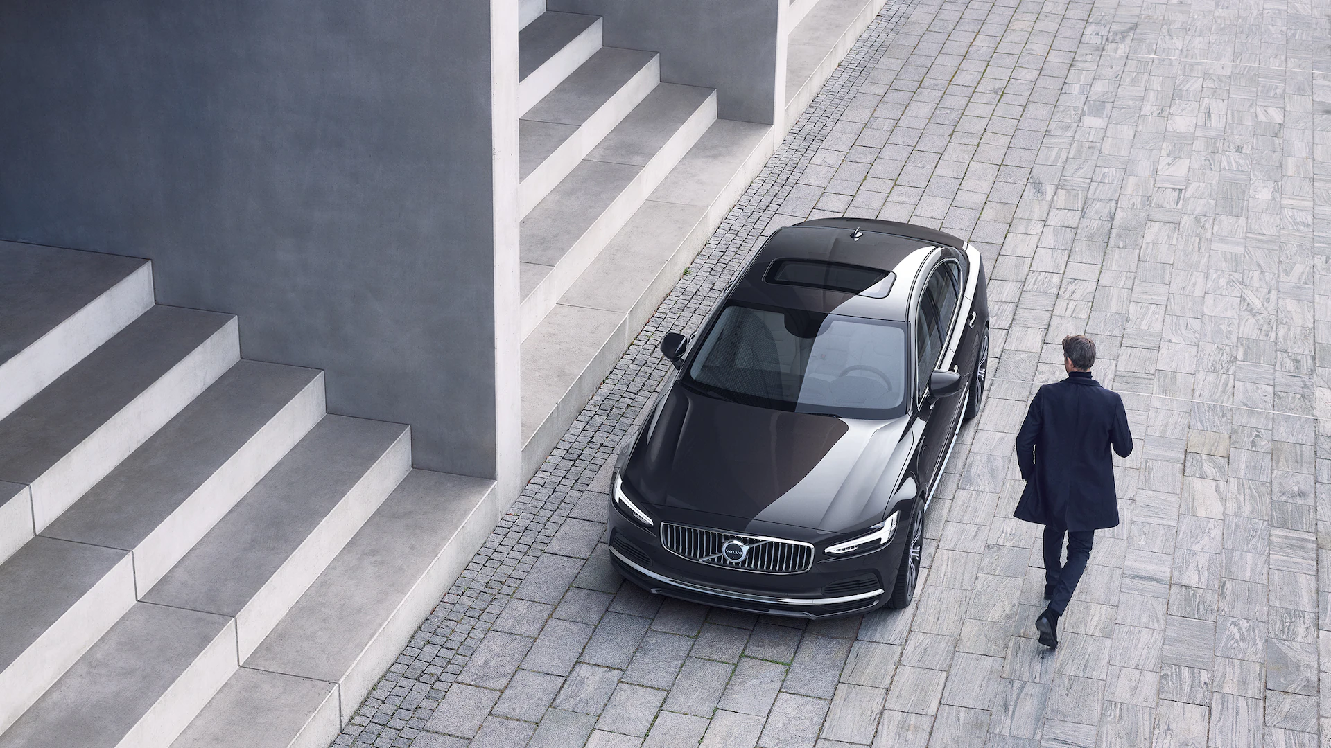 A Volvo S90 is parked in front of a set of stairs, a man walks towards the car.