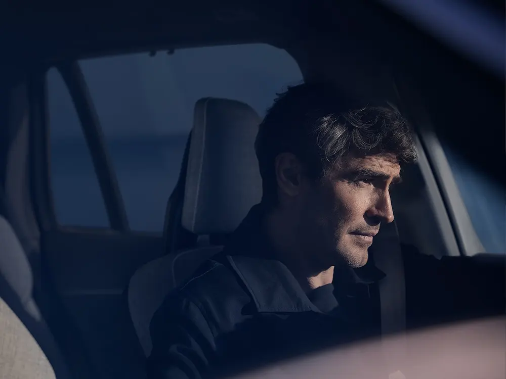 A brown-haired man sits in a Volvo XC90 Recharge and gets the sun in his face.