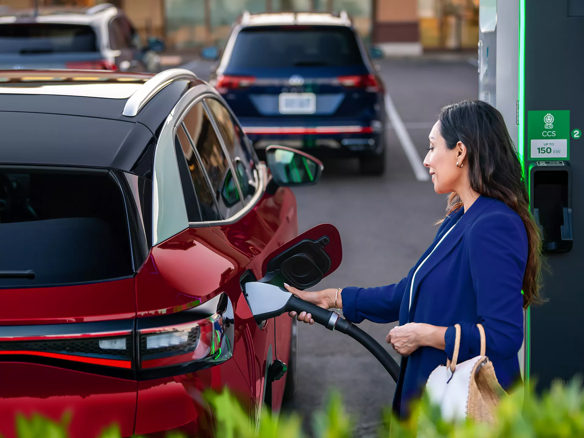 A well-dressed woman, parked at charging station stands on the passenger side of an ID.4 SUV in Aurora Red Metallic plugging a charging cable into the vehicle.