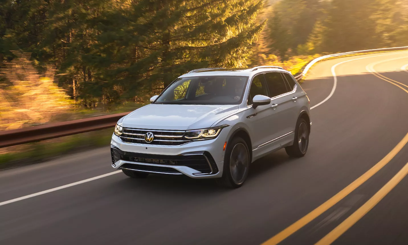 A Tiguan shown in Opal White drives down a winding, two-lane wooded road, as seen from the front driver side.