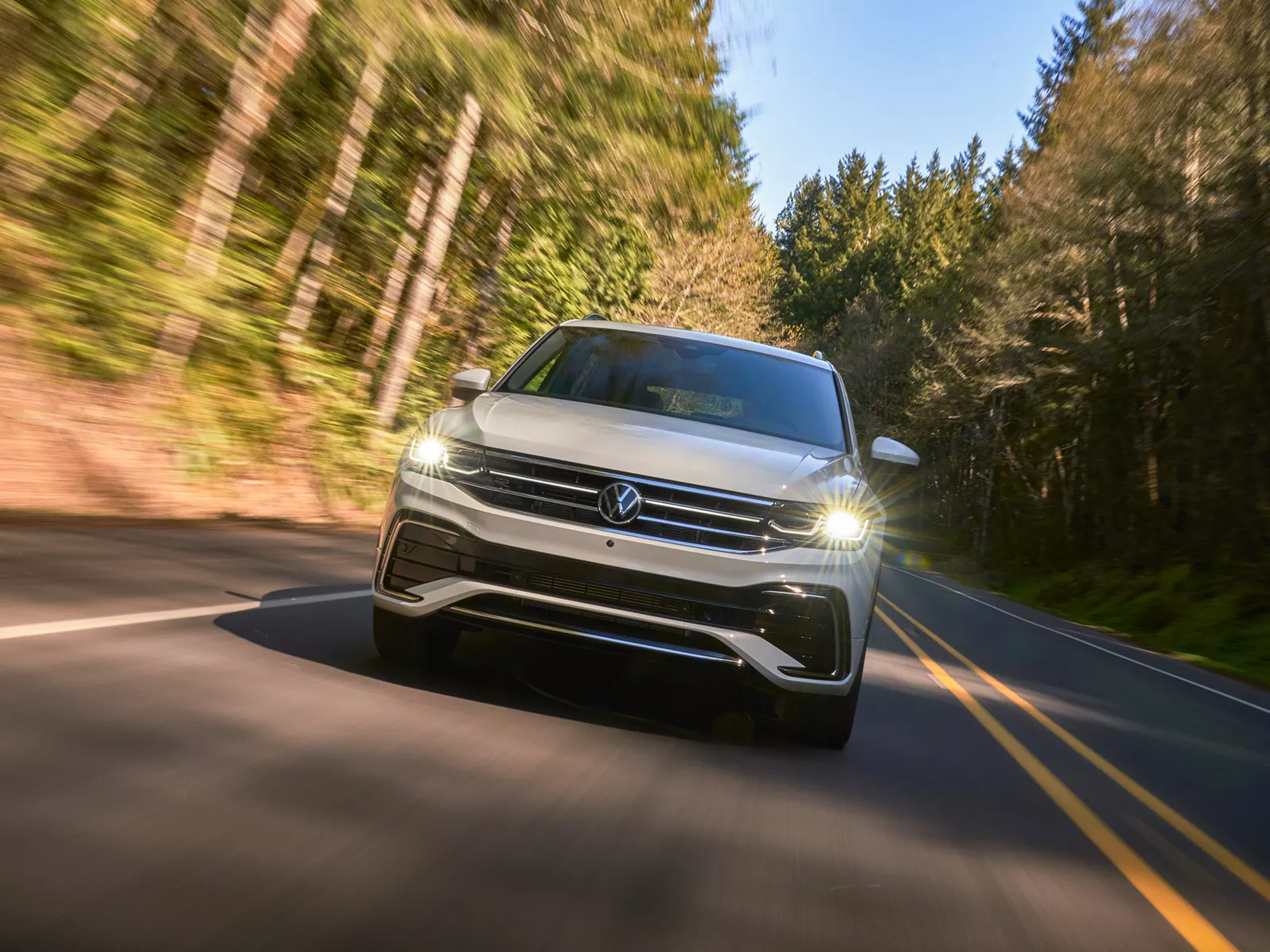 A Tiguan shown in Opal White, driving on a two-lane road with trees in the background, as seen from front.