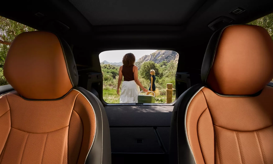 Interior view looking out the open hatch of a parked Tiguan shown in Amber Brown leather with two people holding picnic gear seen outside the vehicle in a wooded park setting.